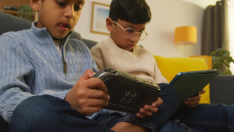 Two-Young-Boys-Sitting-On-Sofa-At-Home-Playing-Games-Or-Streaming-Onto-Digital-Tablet-And-Handheld-Gaming-Device-2